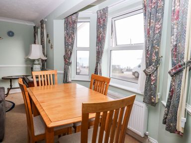 A dining area with a wooden table and chairs next to windows with floral curtains at Ellan Vannin in Sheringham