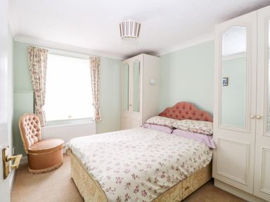 A bedroom with a bed with floral bedding a pink upholstered chair two wardrobes and a window with curtains at Ellan Vannin in Sheringham