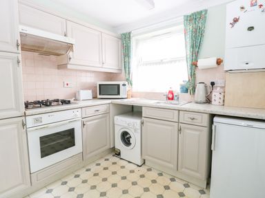 A kitchen with white cabinets a gas stove an oven a microwave a washing machine a refrigerator a kettle and a window with floral curtains at Ellan Vannin in Sheringham