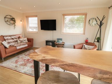 A living room with a brown sofa a wooden table a wall-mounted television and a clock at 3 Valley View Lodges Nawton near Helmsley