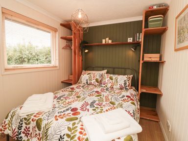 A bedroom with a bed covered in leaf patterned bedding two folded towels on the bed wooden shelves around the bed a window with blinds and a framed picture on the wall at 3 Valley View Lodges in Nawton near Helmsley