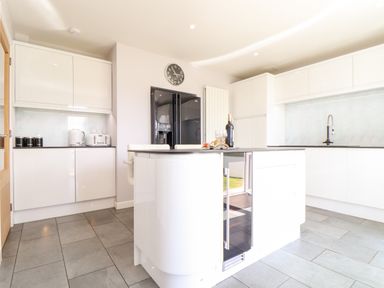 A kitchen with white cabinets a black refrigerator an island with a wine bottle and glasses and a wall clock at Summerhayes in Park Bottom