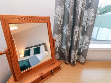 A wooden framed mirror on a wooden desk reflecting a bed with green cushions and a lamp with patterned curtains beside a window at Summerhayes in Park Bottom