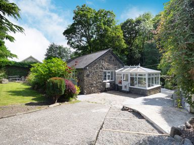 A stone cottage with a glass conservatory surrounded by trees and garden at Nant Cottage in Rowen
