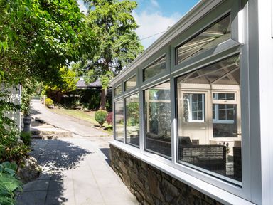 A paved pathway beside a conservatory with glass windows and garden with trees and shrubs at Nant Cottage in Rowen