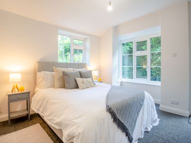 A bedroom with a bed pillows bedside tables with lamps and windows showing greenery at Nant Cottage in Rowen