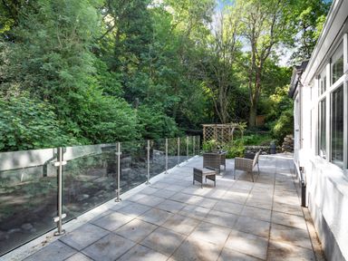 An outdoor patio with glass railing and wicker furniture next to a white building surrounded by trees at Nant Cottage in Rowen