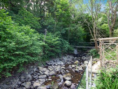 A rocky stream flowing through a forest with green trees and a metal pedestrian bridge in Rowen