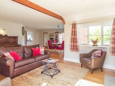 A living room with a brown leather sofa and armchair red and plaid cushions beside a dining area with red chairs and a window with plaid curtains at The Croft in Thompson near Watton