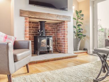 A living room with a brick fireplace and wood stove a leather armchair with a cushion a large potted plant a glass door and a rug on a wooden floor at The Croft in Thompson near Watton
