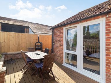 An outdoor wooden deck with a dining table and chairs next to a brick building with white-framed glass doors at The Croft in Thompson near Watton