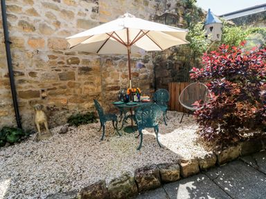 An outdoor patio area with metal chairs and table under a white umbrella a decorative dog statue and a birdhouse on a stone wall at Beals Yard in Alnwick