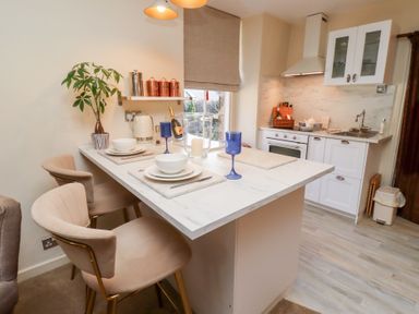 A kitchen area with white cabinets a stove and a sink next to a dining table set for two with blue glasses and beige chairs at Beals Yard in Alnwick