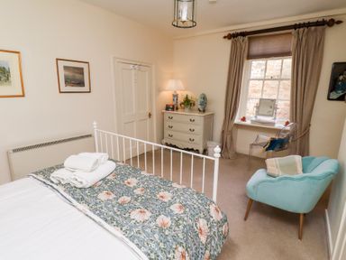 A bedroom with a bed towels and floral blanket a white chest of drawers chair and window with curtains at Beals Yard in Alnwick
