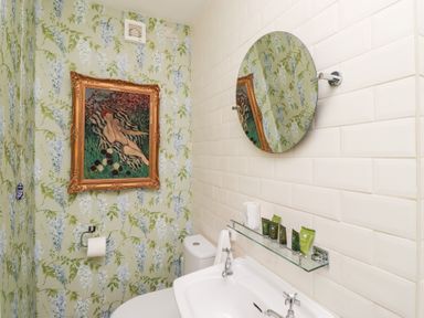 A bathroom with floral wallpaper a round mirror a framed painting a toilet and a sink with toiletries at Beals Yard in Alnwick
