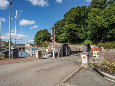 A road leading to a swing bridge over a canal with boats moored alongside and trees in the background at Gwel Y Fenai in Y Felinheli