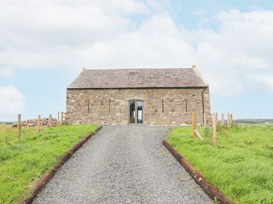 A gravel driveway leading to a stone building with a slate roof and an arched door surrounded by green grass at Gwel Y Fenai in Y Felinheli