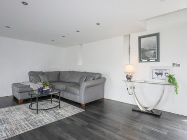 A living room with a grey sectional sofa a glass coffee table with a teapot and cups a patterned rug a glass side table with a lamp framed pictures and a plant at Gwel Y Fenai in Y Felinheli