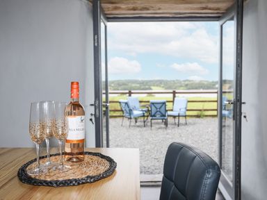 A table with a bottle of wine and four glasses in front of open glass doors leading to outdoor chairs and a scenic view at Gwel Y Fenai in Y Felinheli