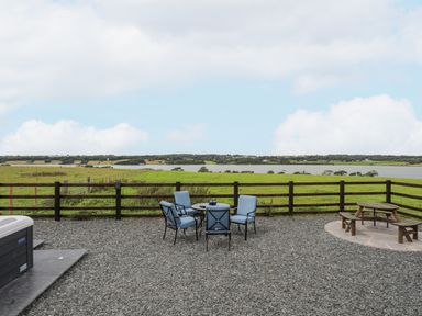 An outdoor patio with blue cushioned chairs around a round table a wooden picnic table and a hot tub overlooking green fields and a body of water at Gwel Y Fenai in Y Felinheli