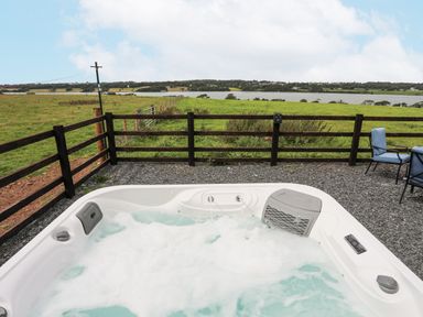 An outdoor hot tub with water jets and foam on a gravel surface near a wooden fence overlooking green fields and water at Gwel Y Fenai in Y Felinheli