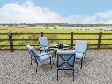 An outdoor seating area with four chairs around a round table on a gravel surface with a wooden fence and green fields and water in the background at Gwel Y Fenai in Y Felinheli