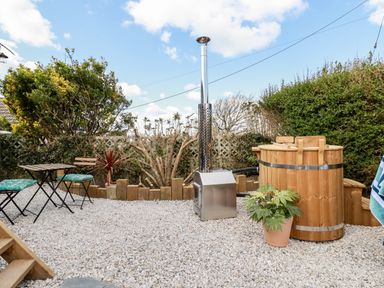 An outdoor patio area with a wooden hot tub a metal stove two chairs with cushions and a small table surrounded by bushes and gravel at Badgers Rest in Tintagel
