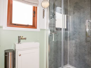 A bathroom with a small white sink cabinet under a wooden window and a glass shower enclosure with grey walls at Badgers Rest in Tintagel