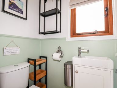 A bathroom with a toilet, a small sink, a window, a toilet paper holder shaped like a knight, a wooden shelf unit, and a wall sign at Badgers Rest in Tintagel