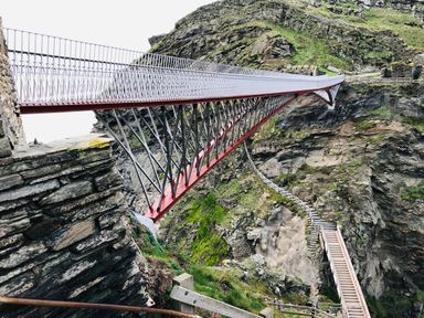 A metal suspension bridge over rocky terrain with stone walls and wooden stairs at Badgers Rest in Tintagel