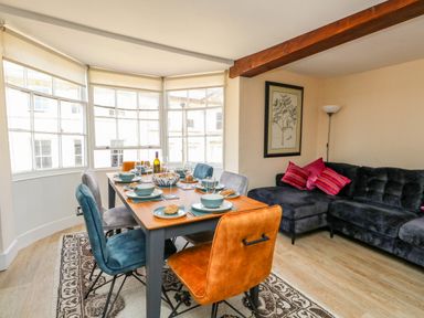 A dining area with a wooden table set for six and a blue and orange chairs next to a dark grey sectional sofa with pink cushions at The Town House in Dorchester