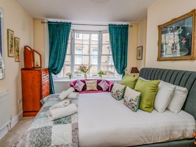 A bedroom with a large bed green pillows a pink sofa by a bay window with blue curtains and a wooden dresser at The Town House in Dorchester