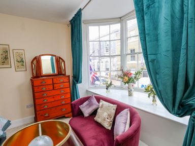 A room with a wooden dresser with mirror a purple sofa with cushions teal curtains and a window sill with flower vases at The Town House in Dorchester