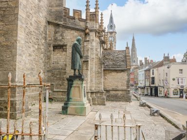 A stone statue on a pedestal beside a historic building with a clock tower and spires in the background at The Town House in Dorchester