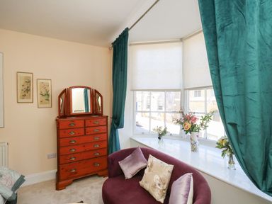 A bedroom with a wooden dresser with mirror green curtains a purple chair and flowers on a window ledge at The Town House in Dorchester