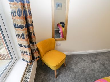 A yellow chair near a window with patterned curtains and a wall mirror reflecting part of a bed at Harbour Hideaway in Whitby