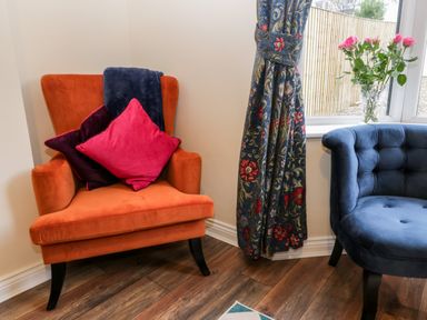 An orange armchair with cushions and a blue throw next to a blue armchair by a window with floral curtains and a vase of pink flowers at Harbour Hideaway Whitby
