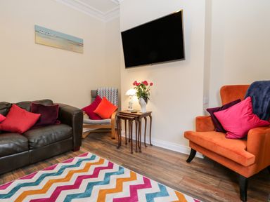 A living room with a brown leather sofa red and purple cushions a wooden armchair with red and orange cushions an orange armchair with purple and red cushions a chevron patterned rug a wall mounted tv and nesting tables with a lamp and flowers at Harbour Hideaway in Whitby