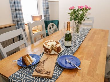 A dining table with a cake a bottle of champagne two blue plates with forks and a vase of pink roses at Harbour Hideaway Whitby