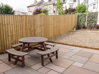 An outdoor patio area with a round wooden picnic table with benches beside a gravel section and a wooden fence at Harbour Hideaway Whitby