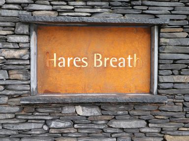 A rust-colored metal sign with the text Hares Breath mounted on a stone wall at Hares Breath in Bowness-On-Windermere