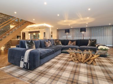 A living room with a large blue sectional sofa a glass coffee table with wooden base plaid rug and staircase at Hares Breath in Bowness-On-Windermere