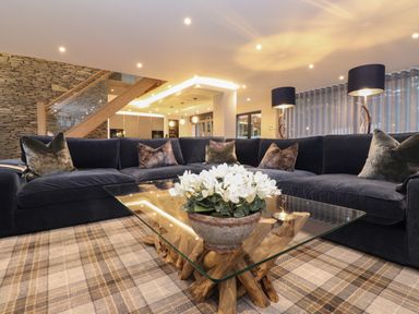 A living room with a large dark blue sofa a glass coffee table with white flowers and two floor lamps at Hares Breath in Bowness-On-Windermere