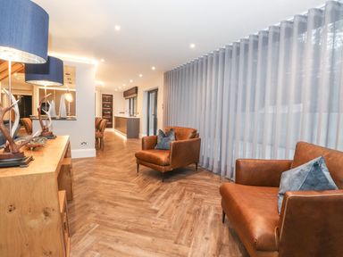A living area with two brown armchairs with blue cushions wooden sideboard with lamps and antler decor and sheer curtains at Hares Breath in Bowness-On-Windermere