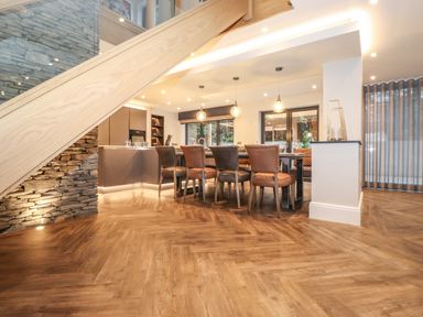 A dining area with a table and six chairs under pendant lights adjacent to a kitchen and stairs with a stone wall at Hares Breath in Bowness-On-Windermere