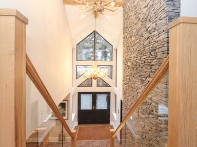 A staircase with wooden and glass railings leading down to a front door with multiple windows and a stone wall on one side at Hares Breath in Bowness-On-Windermere
