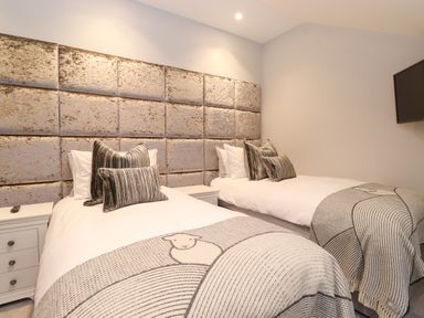 A bedroom with two single beds with cushions and throws a wall-mounted TV and bedside tables at Hares Breath in Bowness-On-Windermere
