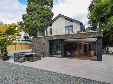 An outdoor seating area with table and benches on a tiled patio in front of a modern two-story house with stone and glass exterior at Hare's Breath in Bowness-On-Windermere