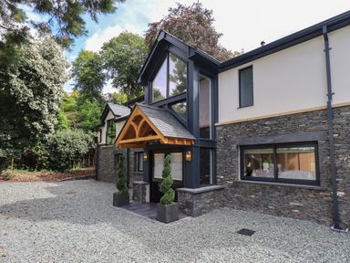 The exterior of a modern house with a stone facade large windows a wooden entrance canopy and gravel driveway surrounded by trees at Hares Breath in Bowness-On-Windermere