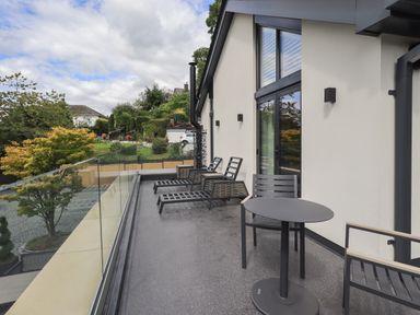 An outdoor balcony with black lounge chairs and a small round table overlooking a garden at Hares Breath in Bowness-On-Windermere
